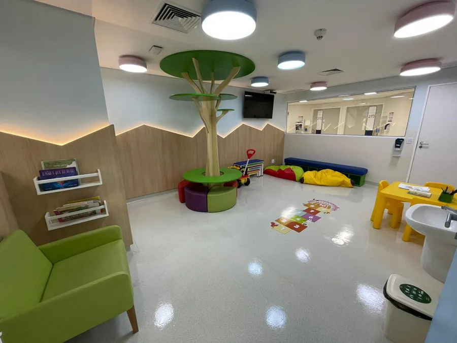 Brinquedoteca de hospital com árvore central de assentos, poltrona para acompanhante, mesa infantil, amarelinha no piso, lavatório e visor de vidro para o corredor da internação